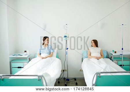 Two pregnant women resting in beds in hospital room and talking about children upbringing