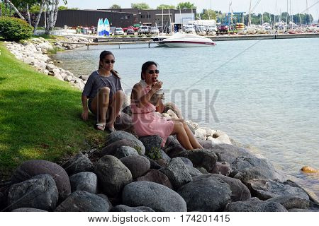 HARBOR SPRINGS, MICHIGAN / UNITED STATES - AUGUST 4, 2016: A woman eats Kilwin's chocolate ice cream, while sitting on a rock along the Harbor Springs waterfront, and enjoying the view of Little Traverse Bay.