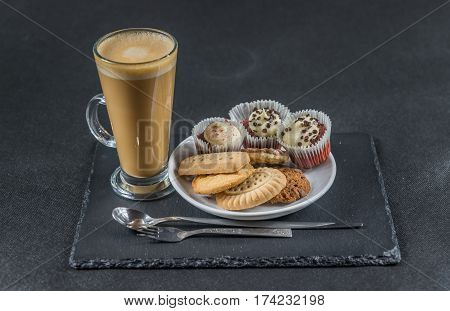 aromatic coffee latte with three buttered muffins and petit fours on a white saucer with a spoon and forks the whole on a black stone plate sweet set