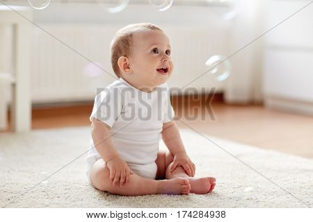 childhood, babyhood and people concept - happy little baby boy or girl sitting on floor with soap bubbles around at home