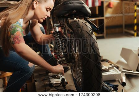 Work hard. Responsible girl using wrench while fixing damper to motorbike, sitting in profile in the workshop