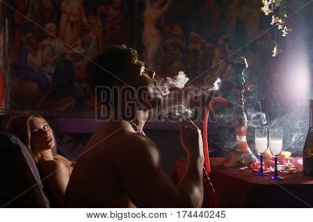 Young handsome athletic tattooed man smoking hookan with gorgeous blonde in bicini sitting on the background shot in luxurious sauna interior