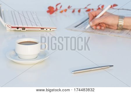 White working Desk with Business Items portable Laptop pen Coffee Mug and red Flower Hand of Person making Notes in Morning Newspaper