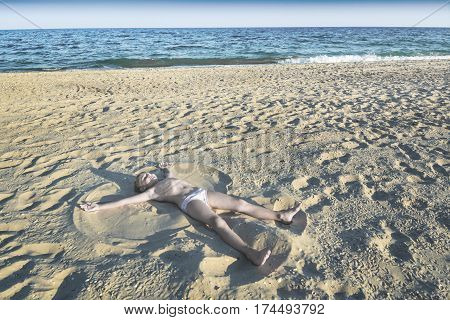 Little boy playing on sea beach making an angel on yellow sand.