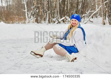 Plump smiling woman in skates sits in snowdrift at outdoor skating rink in winter park.