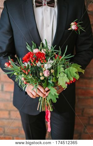 the elegant man dressed in a modern black suit and white shirt holding in hand a wedding flowers bouquet from red carnation. Close up color vertical image of male hands holding bunch of bright flowers.