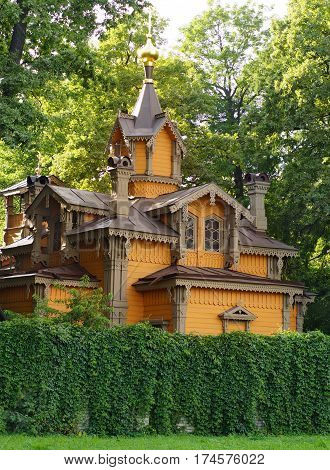 Church of orange color of wood with carved details is behind the green fence surrounded by trees.