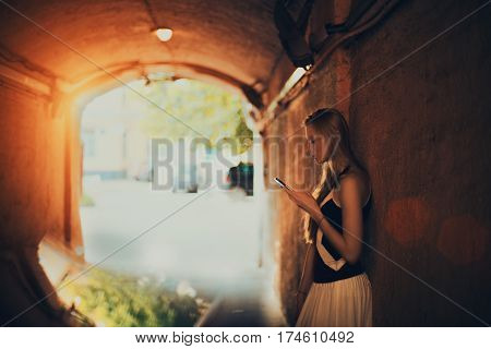 Adult blonde woman surfing internet via smart phone while standing in dark street tunnel and waiting her friend flirty lady is chatting with boyfriend using cell telephone on sunny summer day