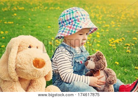 sad boy sitting with softtoys outoors. Thoughtful child with teddy and plush dog on the field with dandelions in spring