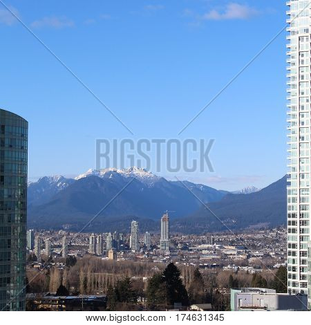 City landscape view on sunny day with high rise buildings and trees in foreground. Snow covered mountain peaks and clear blue sky in background. Burnaby BC Canada.