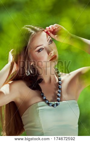 Close-up portrait of a beautiful young woman with your hands near the headlong hair a red lips a thoughtful gaze at the open air in the middle of green blurred leaves at sunny spring day