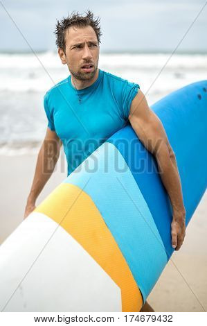 Handsome guy with the disheveled hair and the stubble stands on the beach on the blurry background of the ocean waves and the sky. He wears a cyan T-shirt and holds a colorful surfboard. Closeup.