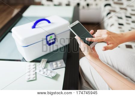 Woman using smartphone for buying drugs in internet pharmacy and getting online medical consultation with doctor at home. White box with first aid kit and pills in blister packs on table.
