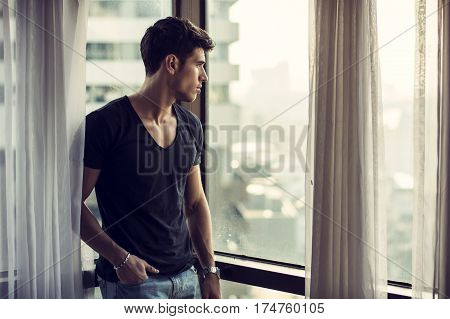 Sexy handsome young man standing next to window curtains, looking out