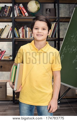 Schoolboy holding books while standing near chalkboard and smiling at camera