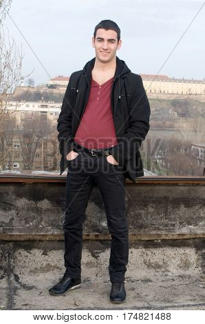 Photographing smiling Danila Mazić in a tight red shirt unbuttoned covered with a black coat and black pants a background Petrovaradin fortress and the Danube River, Novi Sad, Serbia