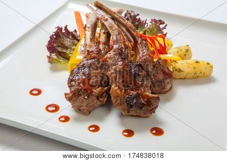 Pork ribs on the grill with roasted vegetables and boiled potatoes on a white plate decorated with fresh lettuce leaves and sweet peppers