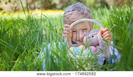smiling happy boy holding basket with easter eggs and bunny toy after easter egg hunt lying in green grass in the park copyspace on the left