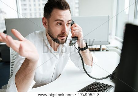 Uncomprehending man in shirt which sitting by the table and talking on phone