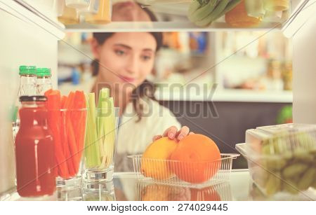Portrait Of Female Standing Near Open Fridge Full Of Healthy Food, Vegetables And Fruits. Portrait O