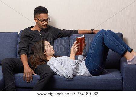 Photo Of Relaxed Caucasian Young Woman Lies On Knees Of Her Dark Skinned Boyfriend, Reads Book Aloud