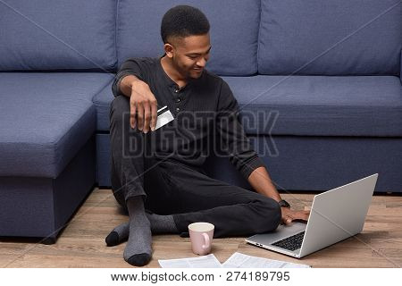 Cheerful African American Guy Holds Credit Card, Checks Her Bank Account On Laptop Computer, Makes O