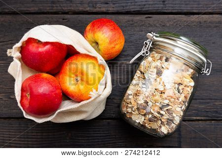 Homemade Granola In Glass Jar And Apples In Reusable Fabric Bag On Dark Wooden Table With Texture. H