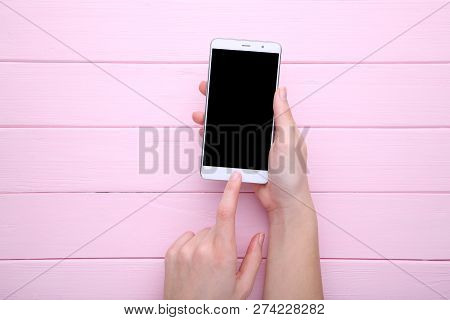 Female Hands Holding Mobile Phone With Blank Screen On Pink Wooden Table. Smartfone White