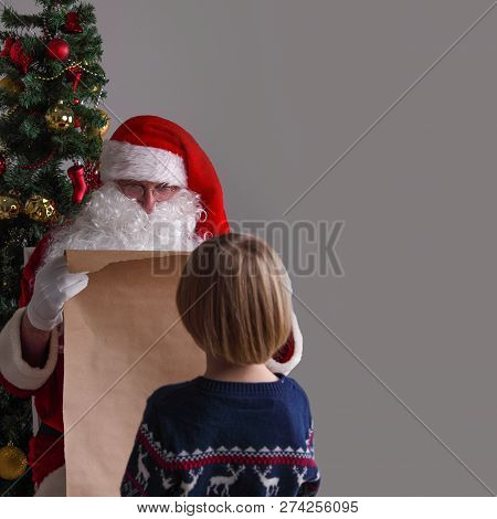 Santa Claus With With List Or Nice Or Naugthy List Paper With Small Boy Near Christmas Tree