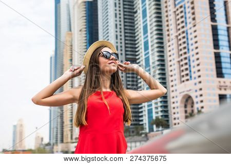 Portrait Of Young Pretty Woman Wearing In Red Dress And Sunglasses, Straw Hat In Front Of Skycrapers