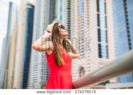 Young Pretty Woman Holding Straw Hat Wearing In Red Dress And Sunglasses In Front Of Skycrapers.