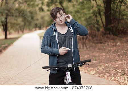 Attractive Young Caucasian Man With Dark Hair Bicycling In The Park. Touches The White Earphones. Ou