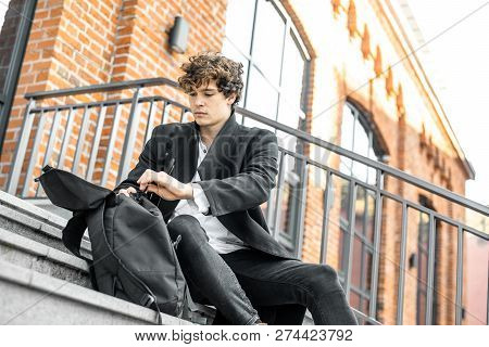 Young Attractive Pensive Man Sitting On Steps And Rummaging In His Backpack.
