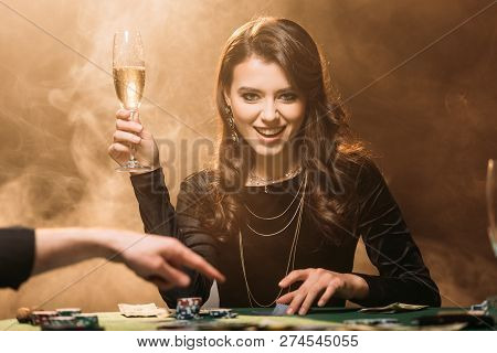 Attractive Happy Girl Holding Glass Of Champagne At Poker Table In Casino And Looking At Camera