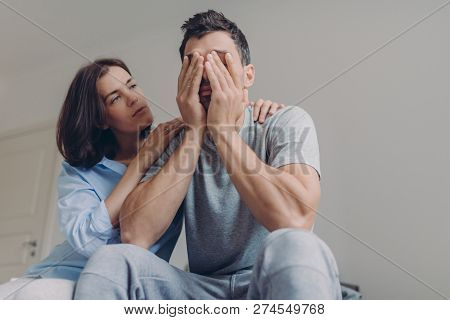 Photo Of Depressed Male Dressed Casually, His Beautiful Woman Comforts Him, Sit Together In Bedroom,