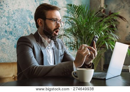 Shocked young businessman dressed in suit working on a laptop computer, sitting at the cafe indoors, holding mobile phone