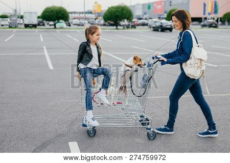 Horizontal Shot Of Happy Woman Carries Rucksack, Dressed In Denim Clothes, Holds Cart, Her Little Da