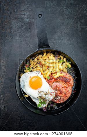 Traditional fried Allgauer veal roast roll sausage slices with fried egg in a cast-iron skillet with spices as closeup on a black board 