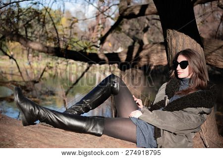 Attractive young woman wearing gray dress and hakki jacket with long fair hair posing outdoor in autunm or spring park over nature outdoor. Beautiful caucasian model sitting on tree near river