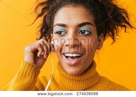African american optimistic woman in casual clothing listening to music via white earphones isolated over yellow background