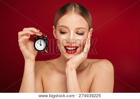 Beauty portrait of a pretty young topless woman wearing makeup isolated over red background, showing small alarm clock