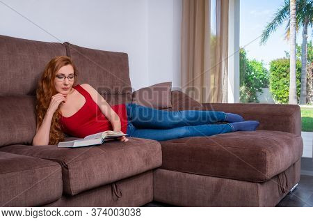 Woman Sitting Comfortably At Her Living Room On A Couch And Reading A Book.