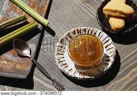 Rhubarb Jam In A Glass Bowl On A Ceramic Plate. There Is A Spoon And Rhubarb Stalks On The Textured 