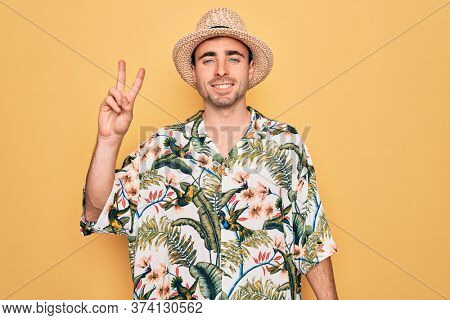 Young handsome man with blue eyes on vacation wearing summer florar shirt and hat smiling with happy face winking at the camera doing victory sign. Number two.