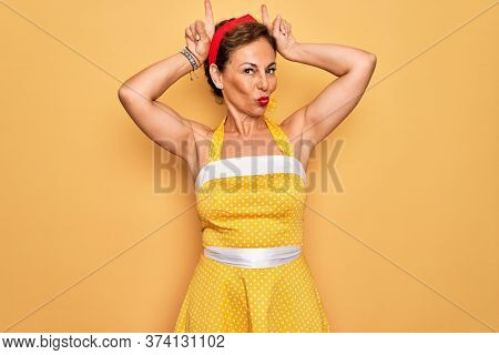 Middle age senior pin up woman wearing 50s style retro dress over yellow background doing funny gesture with finger over head as bull horns