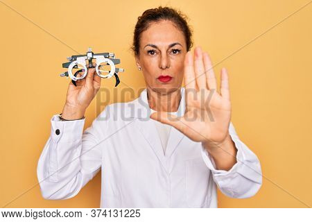 Middle age senior oculist woman holding optometrical glasses for vision correction with open hand doing stop sign with serious and confident expression, defense gesture