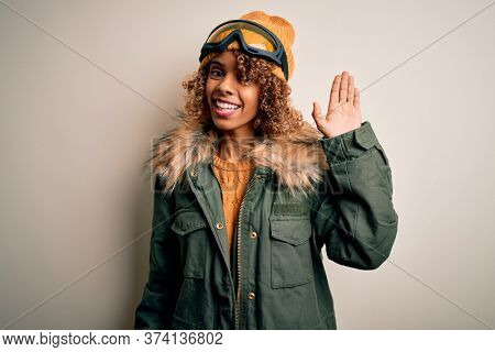 Young african american skier woman with curly hair wearing snow sportswear and ski goggles Waiving saying hello happy and smiling, friendly welcome gesture