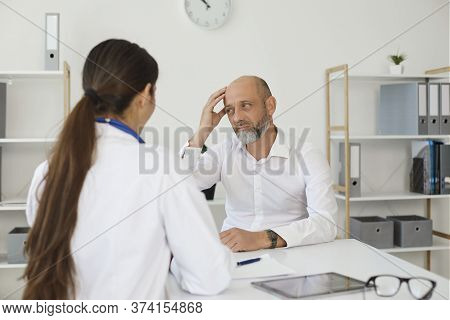 Mature Man With Headache Consulting With Female Doctor At Clinic. Medical Specialist Helping Patient