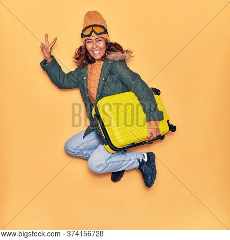 Young beautiful latin woman wearing winter clothes smiling happy. Jumping with smile on face holding cabin bag doing victory sign over isolated yellow background
