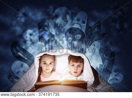 Engrossed Little Kids Reading Book In Bed. Brother And Sister Together Hiding Under Blanket. Fearful